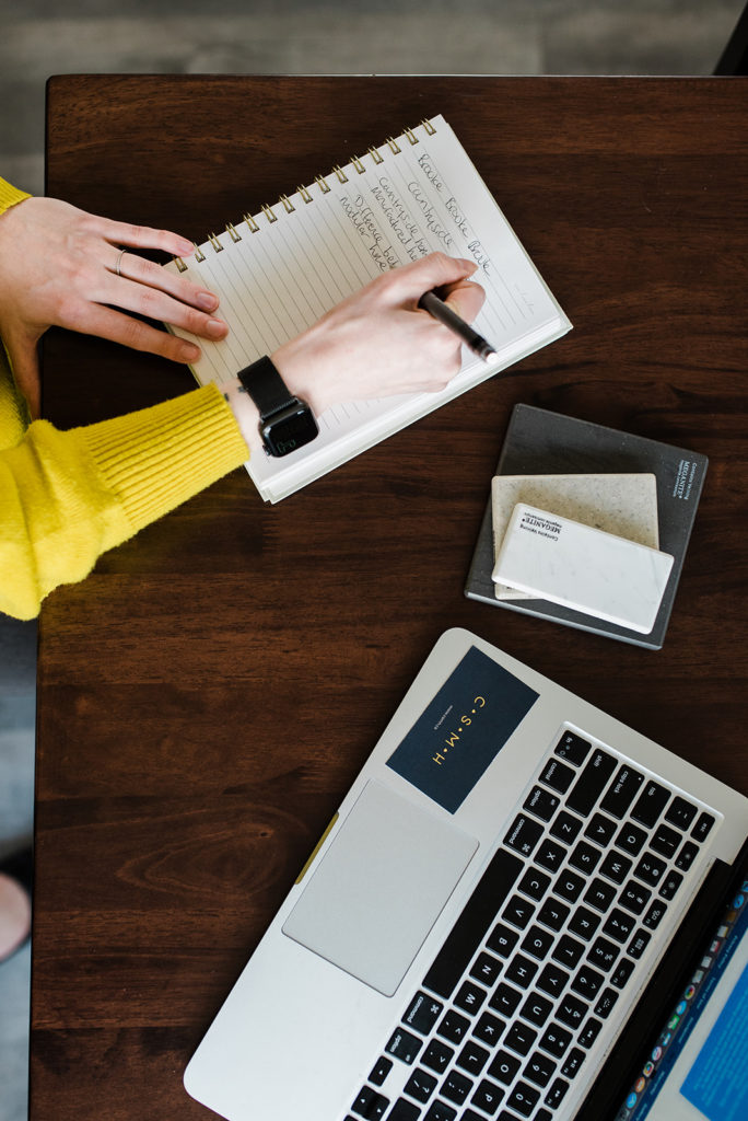 top down of a woman's hands writing in a notebook beside a laptop and business cards